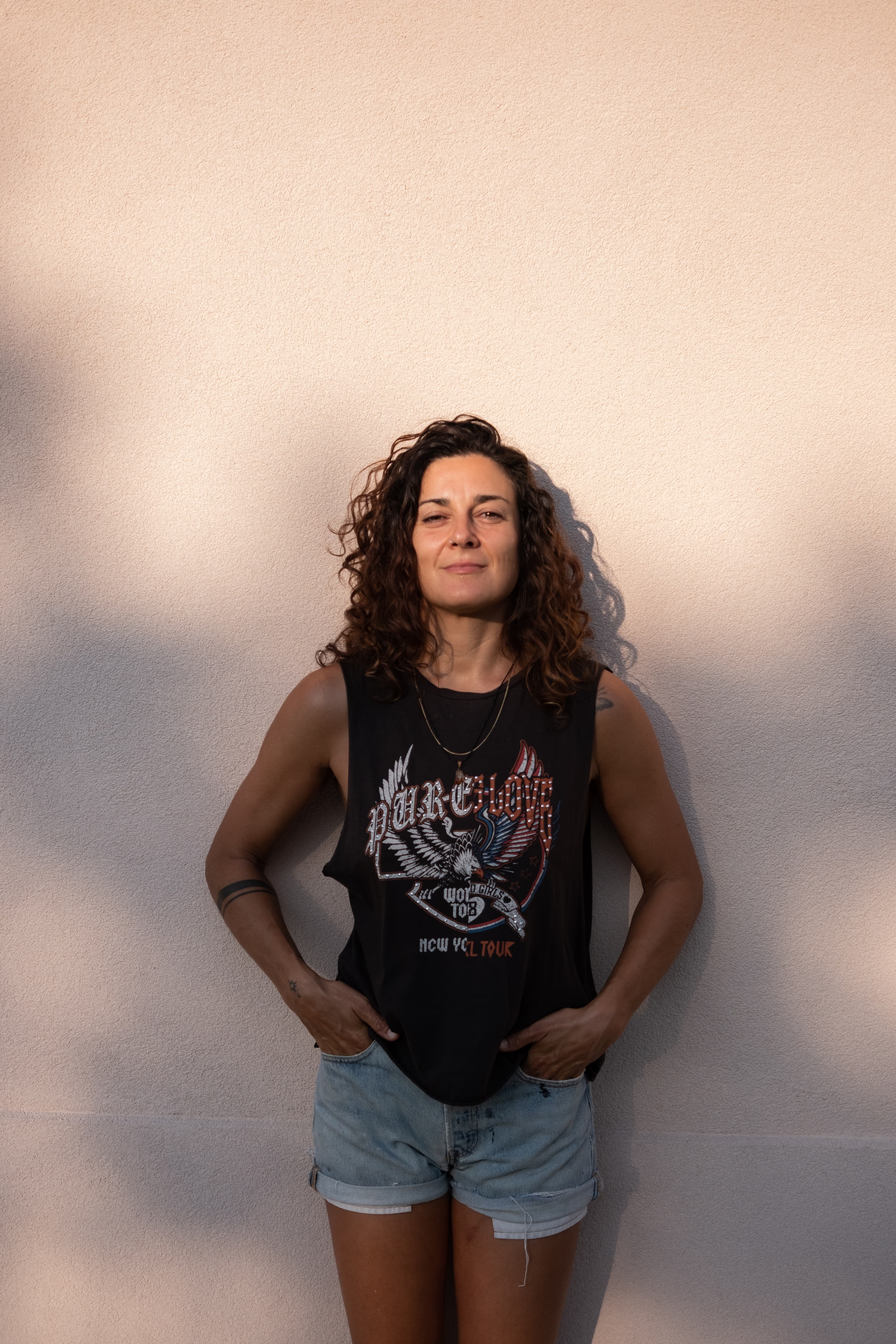 Olivia - Woman with dark curly hair wearing a dark top with 'PURE' graphic design and light denim shorts